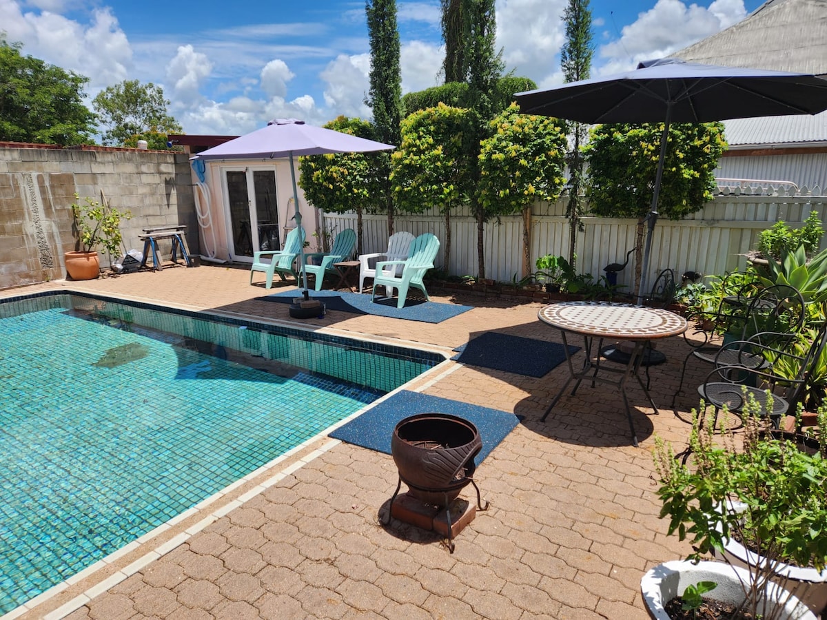 A tranquil pool area is surrounded by lush greenery, featuring two umbrellas providing shade over lounge chairs. A round table with chairs sits nearby, while a fire pit is positioned on the patio. The pool's clear water reflects the bright sky.