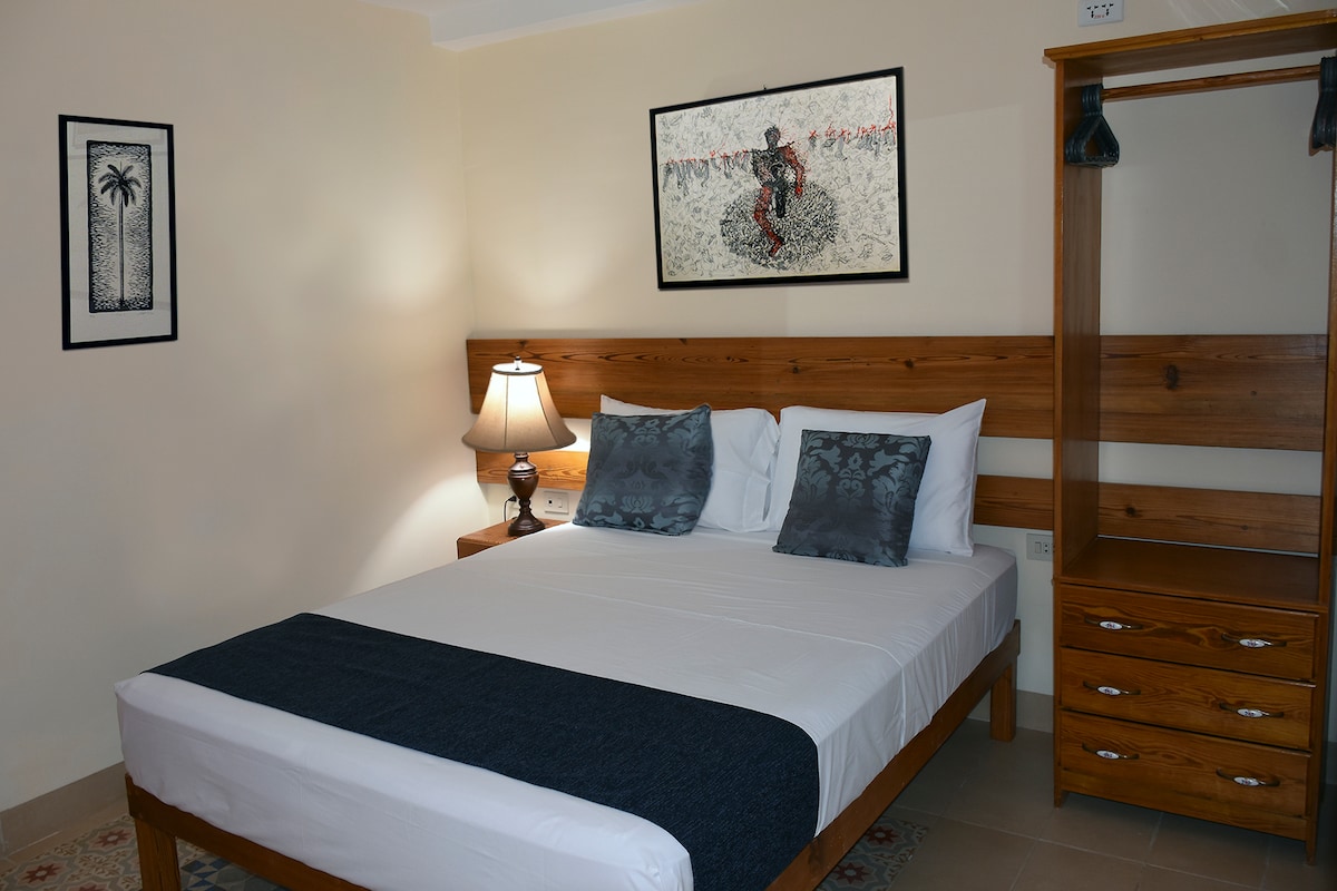 A well-appointed sleeping area features a spacious bed with crisp linens and decorative pillows. A wooden nightstand supports a lamp, providing soft illumination. A wooden wardrobe complements the room, while artwork adorns the wall, adding a touch of character.