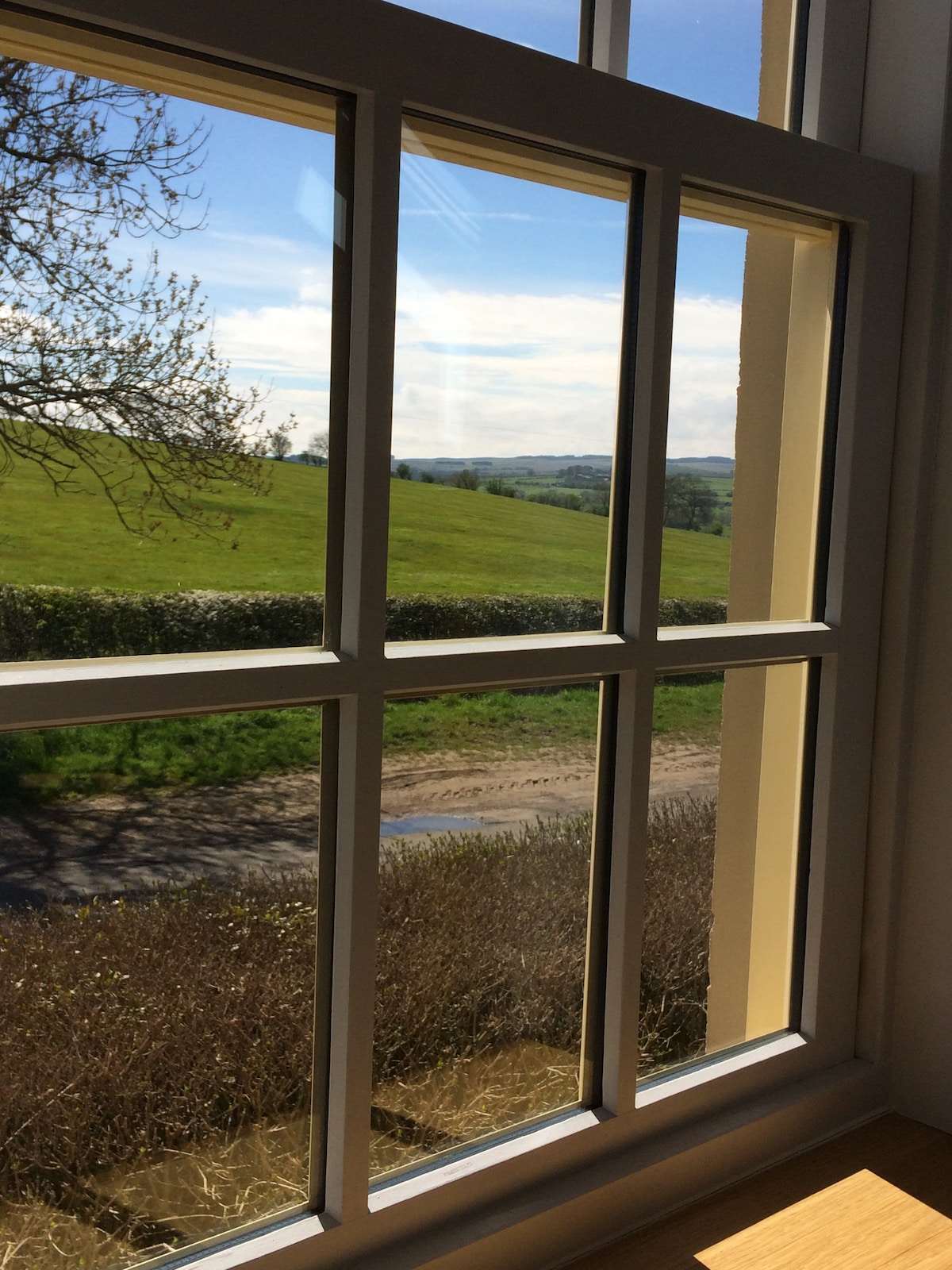 A large window features a six-pane design, offering a view of rolling green fields. The sunlight illuminates the landscape, with a road visible in the foreground. A hedge borders the scene, enhancing the rural setting.