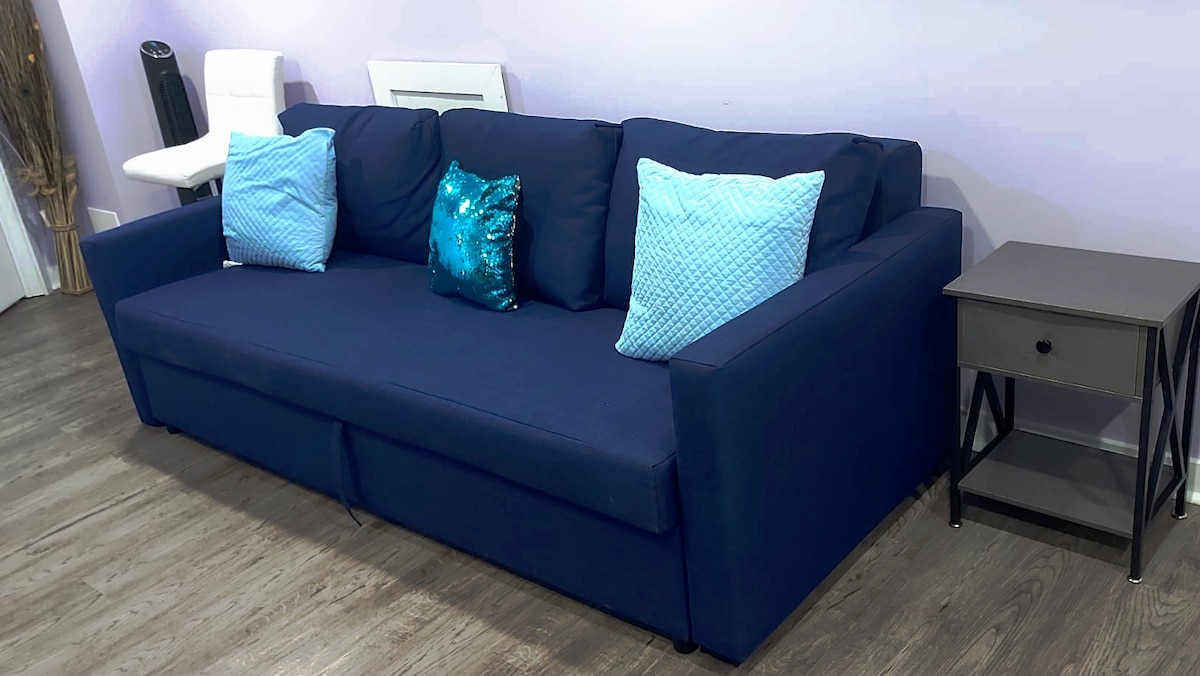 A modern blue sofa with two decorative light blue pillows is positioned against a softly colored wall. A small nightstand, featuring a dark finish, is placed beside the sofa. The flooring is a light wood texture, contributing to the overall welcoming ambiance.