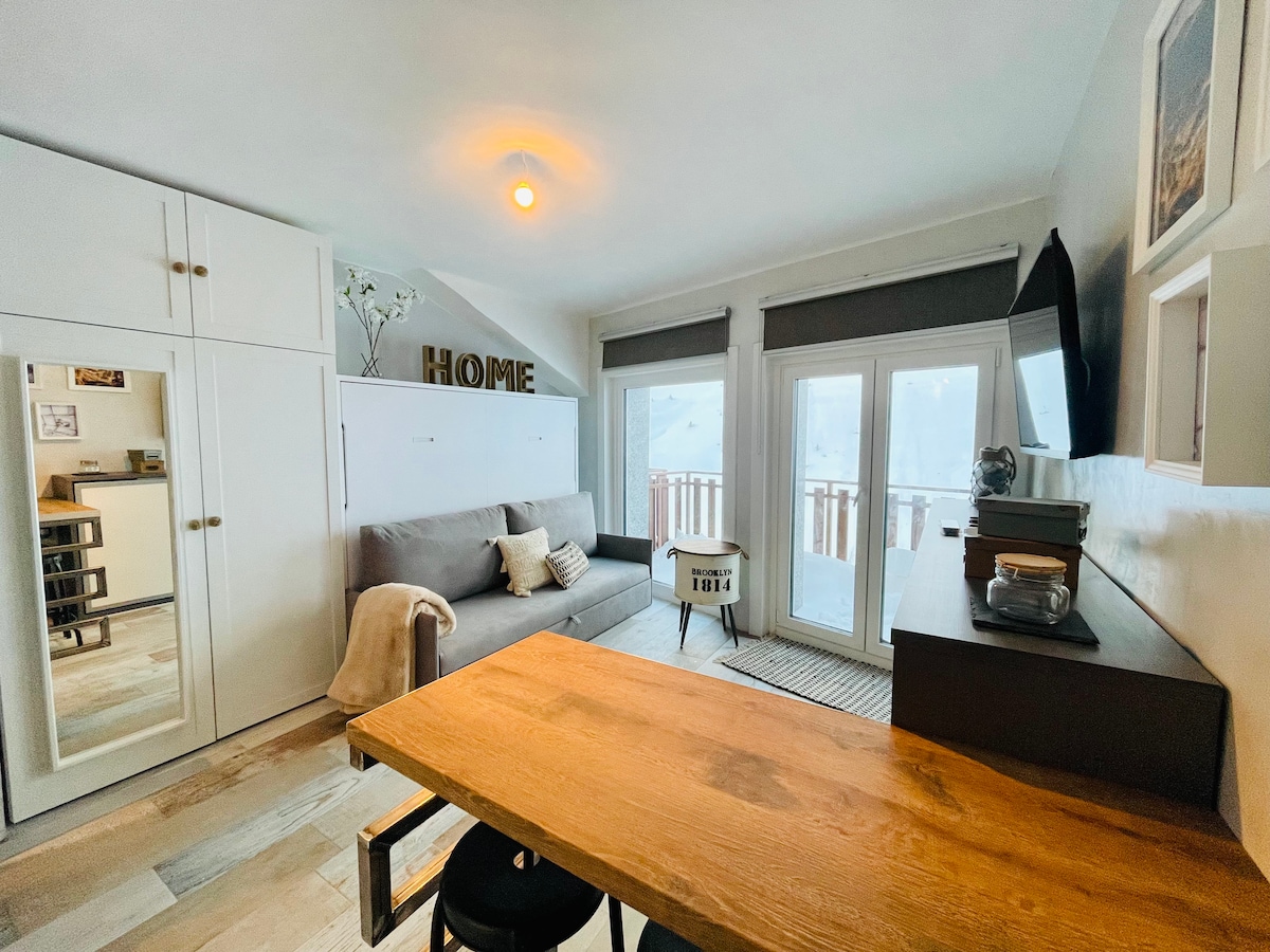 A bright living space features a cozy sofa and a wooden table with stools. A large wardrobe is positioned nearby, and a wall-mounted television is visible. Two large windows provide natural light and access to the balcony, creating a warm and welcoming atmosphere.