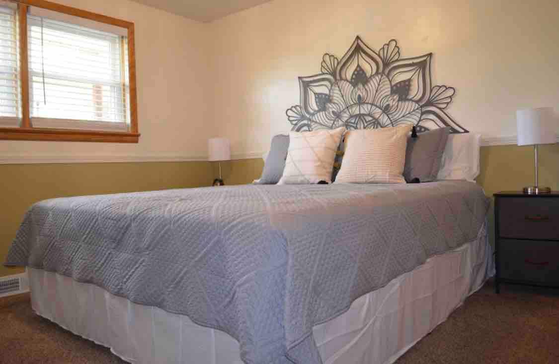 A well-appointed bedroom features a spacious bed with a decorative headboard. Soft neutral tones provide a calming atmosphere. Two bedside lamps are positioned on either side, and natural light filters through a window with horizontal blinds, enhancing the room's welcoming ambiance.
