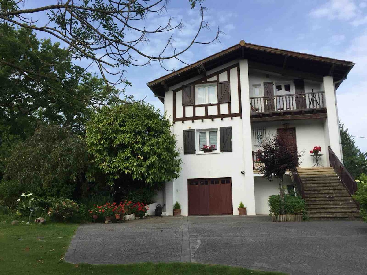 The two-story house features a combination of white stucco and wooden accents. A charming garden surrounds the property, with blooming flowers and lush greenery. A set of stairs leads to the entrance, framed by flower pots, while a balcony overlooks the surrounding area.