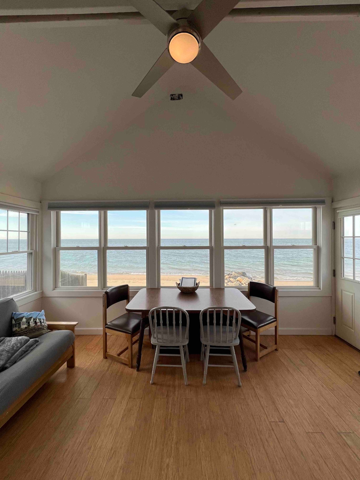 A dining area is featured with a table surrounded by four chairs, positioned near large windows that offer an unobstructed view of Cape Cod Bay. Light hardwood flooring complements the serene atmosphere, and a ceiling fan is visible overhead.