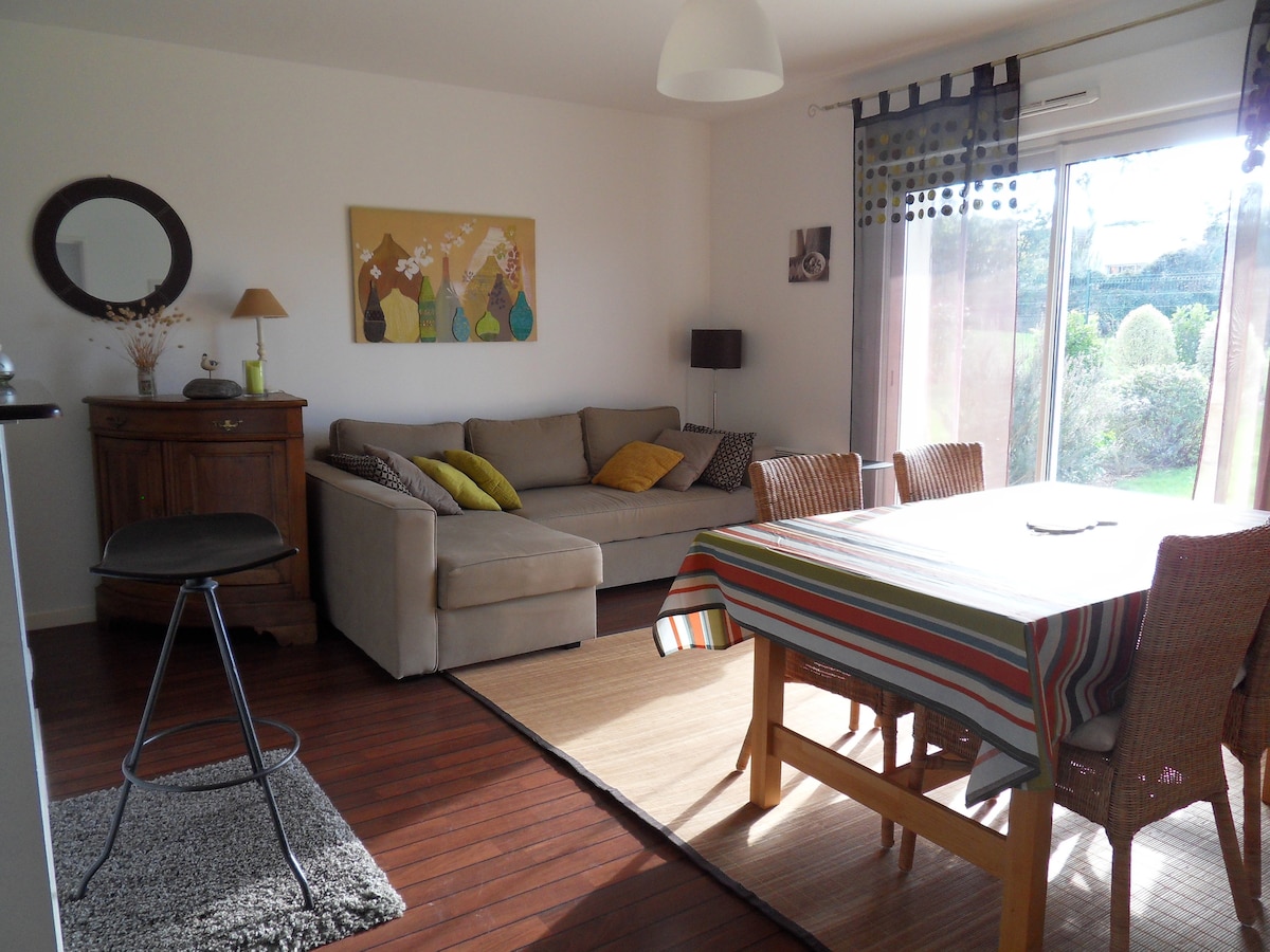 A bright living and dining area features a comfortable sofa adorned with cushions, complemented by a wooden table set with striped tableware. Sunlight filters through large sliding glass doors that open to a garden view, enhancing the open and airy feel of the space.
