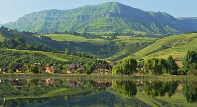 A serene landscape is depicted, featuring rolling green hills and a prominent mountain in the background. A calm water body reflects the scenery, with clusters of rustic buildings nestled among trees in the foreground.