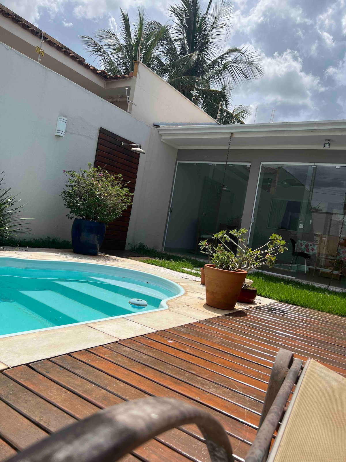 A private pool surrounded by wooden decking offers a relaxing spot, complemented by potted plants and lush green grass. The area is bathed in natural light, with a backdrop of palm trees and a modern glass wall, enhancing the outdoor living space.