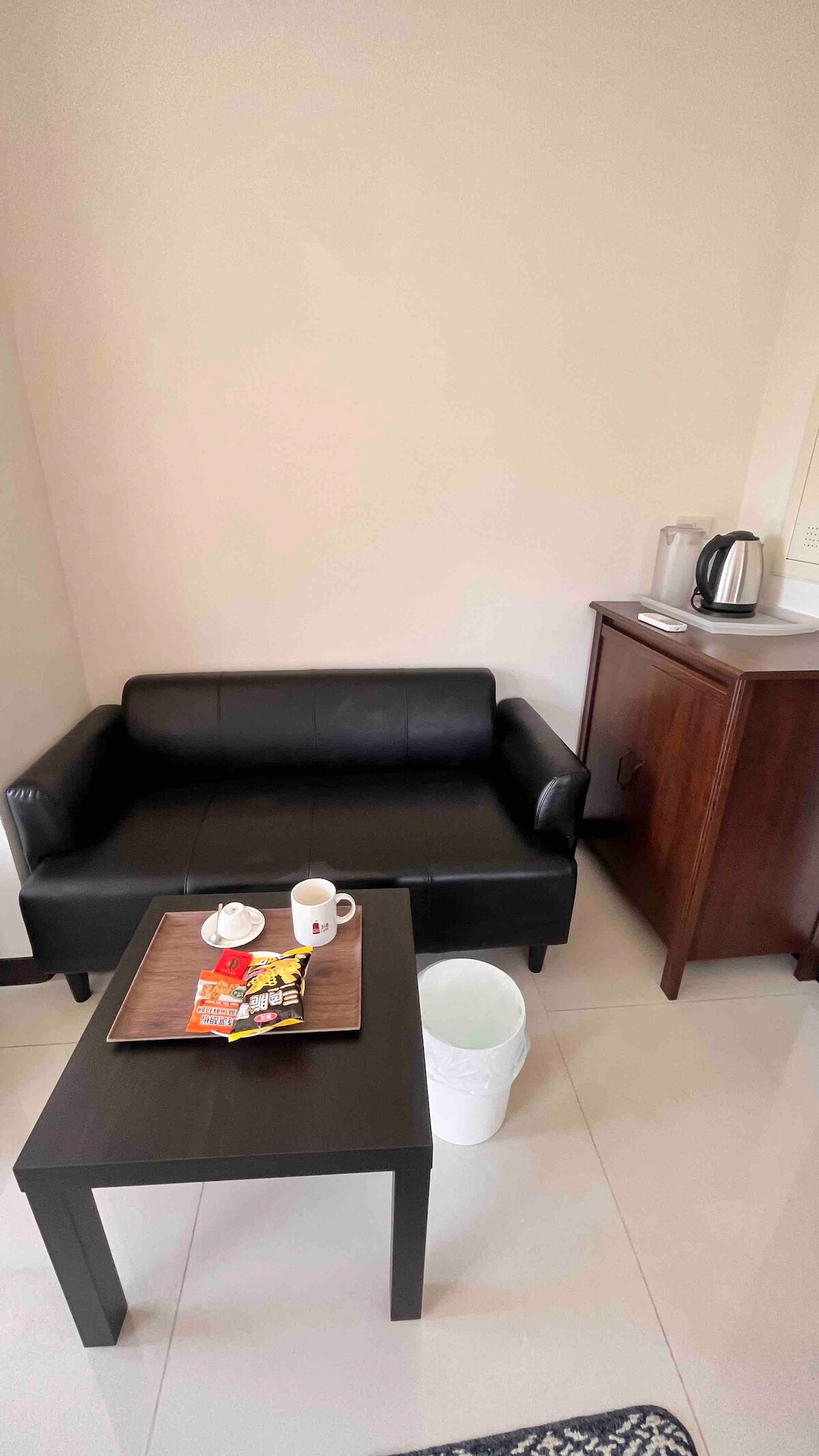 A compact seating area includes a black leather sofa and a small table with snacks and a beverage. A wooden cabinet with a kettle is positioned nearby, enhancing the functionality of the space.