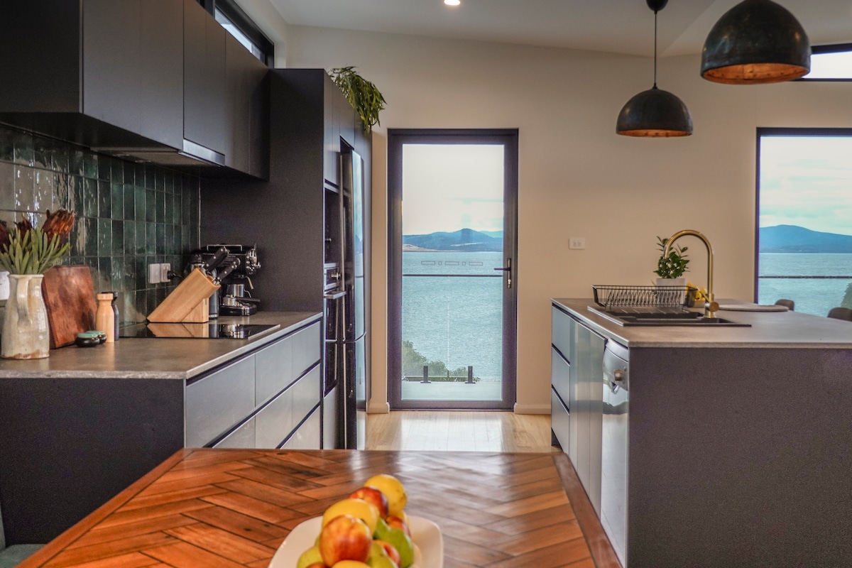 The kitchen features modern cabinetry and stainless steel appliances, complemented by a striking stone backsplash. Natural light flows through large windows, offering views of the water. A wooden dining table holds a bowl of fresh fruit, creating a welcoming space for gatherings.