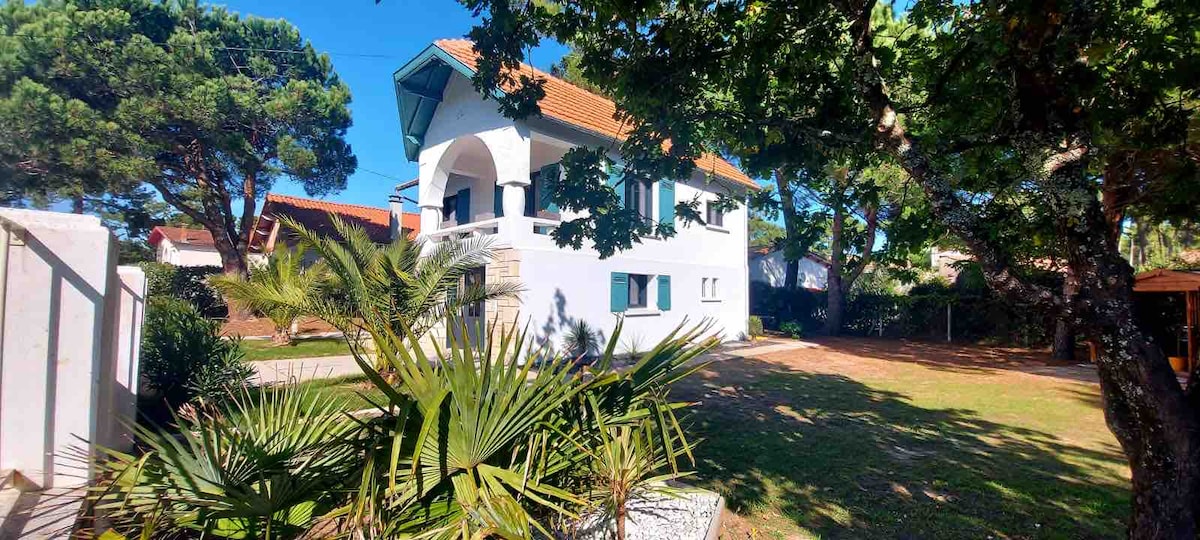 The villa is surrounded by lush greenery, featuring a bright white facade with teal accents. An arched balcony enhances the structure's charm, while large windows allow natural light to fill the interior. A spacious garden offers shaded areas under the trees.