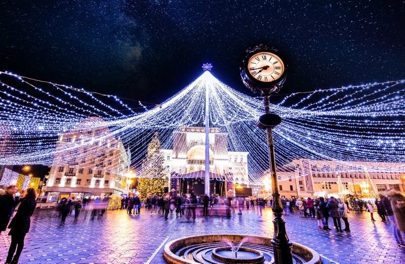 A lively urban square is adorned with twinkling lights overhead, creating a festive atmosphere. A decorative clock stands in the foreground next to a small fountain, surrounded by groups of people enjoying the evening ambiance.