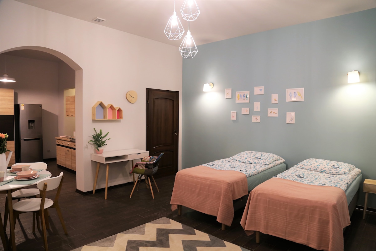 The image showcases a well-lit room featuring two twin beds dressed in light blue and peach linens. A desk with plants and decor is positioned nearby, while a small dining area with tableware is visible to the left. Colorful art pieces adorn the wall, enhancing the space.