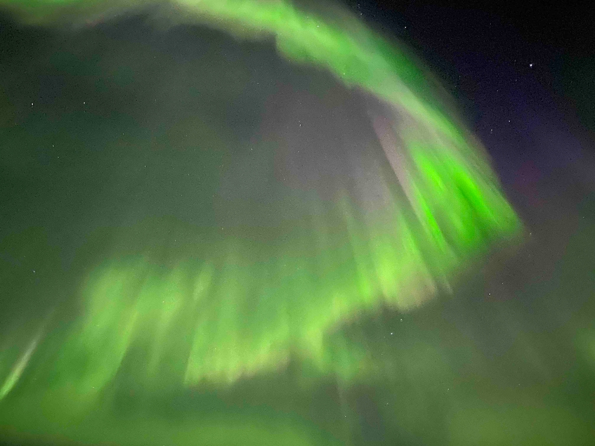The northern lights illuminate the night sky with vibrant shades of green, creating a dynamic display. Wisps of light swirl and dance across the dark background, offering a natural spectacle. Stars are faintly visible in the surrounding sky, complementing the overall scene.