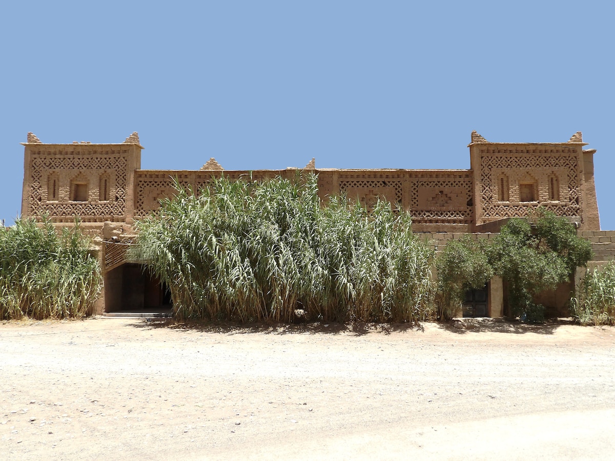 A traditional kasbah is viewed from the front, featuring intricate carvings and a warm, earthen exterior. Lush greenery surrounds the structure, with tall grasses framing the entrance. The sky provides a simple backdrop, highlighting the architectural details of this historic building.