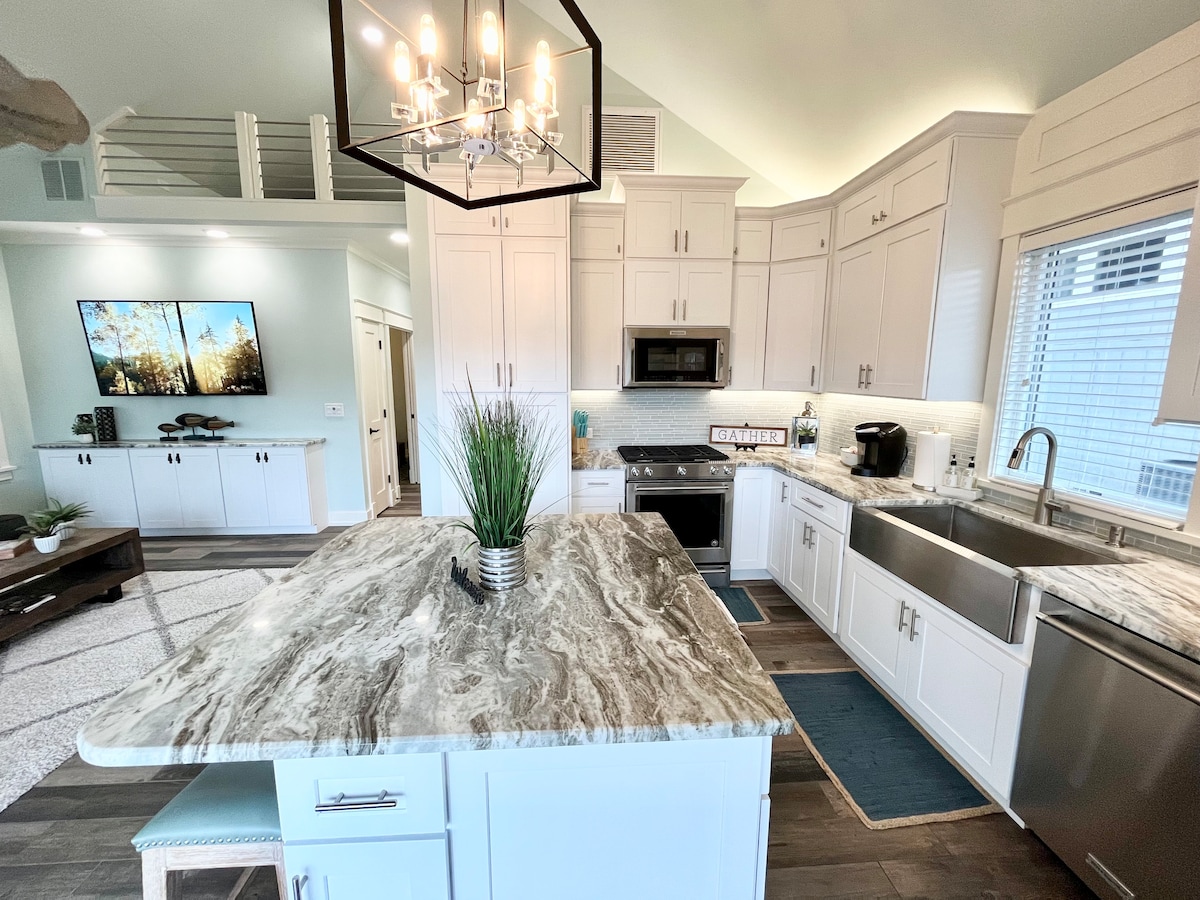 A modern kitchen features white cabinetry and stainless steel appliances, including a microwave and coffee maker. A large central island with a marble countertop provides ample space for meal preparation. Natural light floods the area through various windows, enhancing the clean design.