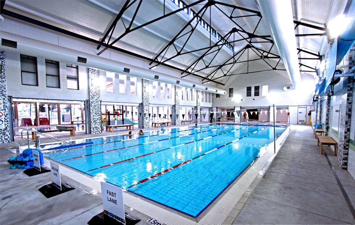 An indoor swimming pool features multiple lanes marked for swimming, with clear blue water reflecting overhead lights. High ceilings and large windows allow ample natural light to fill the space, while benches line the sides for seating.