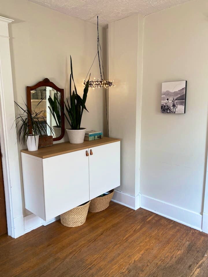 A small entryway features a sleek white cabinet with wood accents, complemented by a mirror and hanging light fixture. Potted greenery adds a touch of nature to the space, while two woven baskets are positioned below the cabinet on the warm-toned hardwood floor.