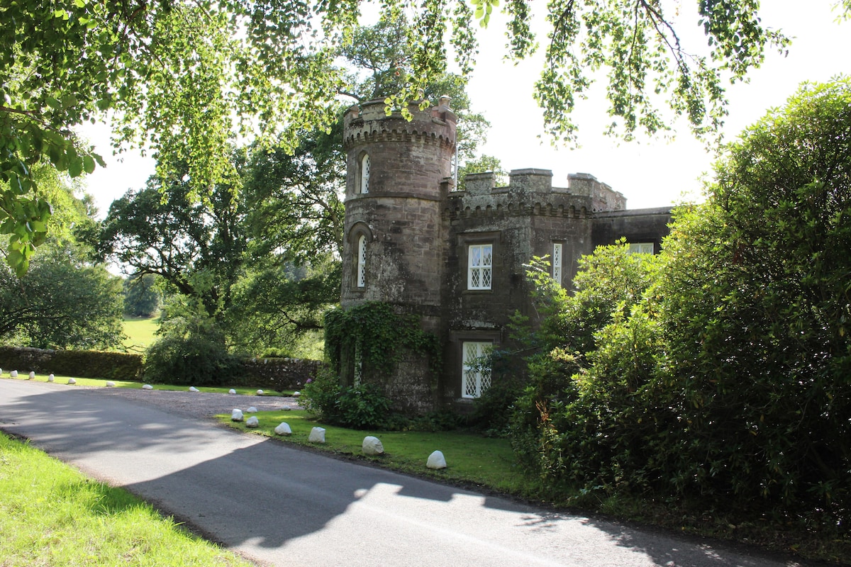 The exterior of the gatehouse is highlighted, showcasing its distinctive turret and stone facade. Lush greenery surrounds the building, with trees and shrubs adding to the natural setting. A quiet road extends in front, offering a serene approach to the property.