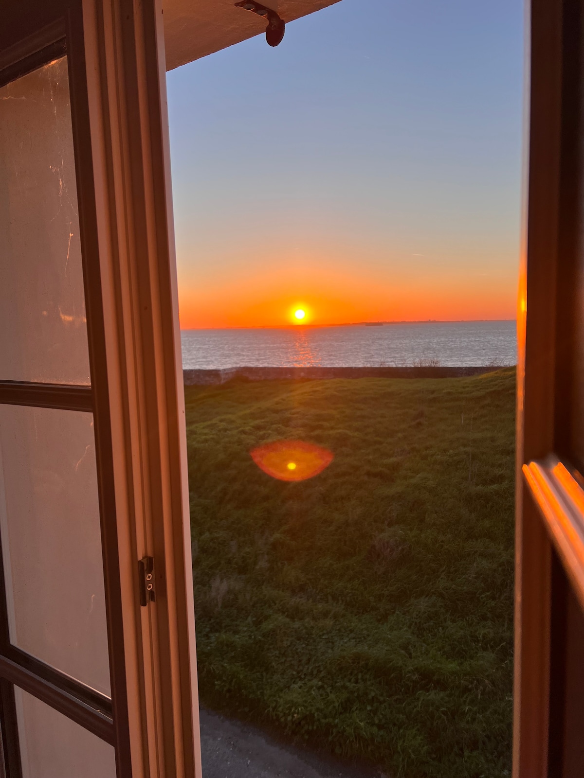 The view from a window captures a vibrant sunset over the sea, with the sun illuminating the horizon. Lush green grass is visible in the foreground, enhancing the serene environment. The window frame adds structure to the scenic landscape.
