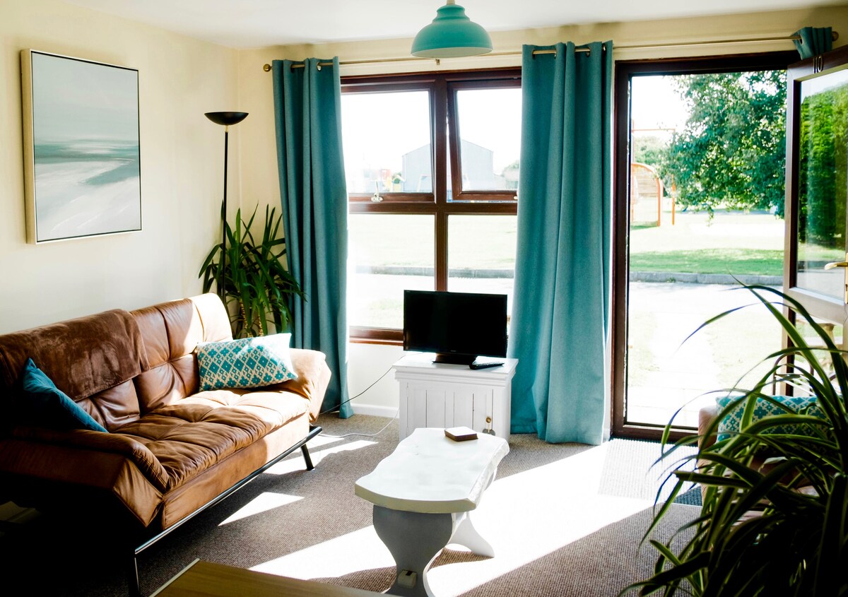 A bright living area is seen, featuring a brown sofa paired with a decorative blue cushion. A light-colored coffee table sits in front of the sofa, with a TV positioned on a white cabinet nearby. Large glass doors open to an outdoor space, framed by turquoise curtains.