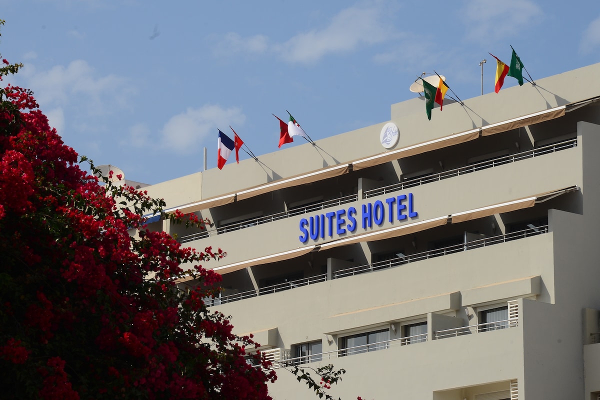 The exterior of the Suites Hotel is visible, featuring multiple flags representing different countries. A vibrant flowering plant in shades of red and pink frames the foreground, contrasting against the light-colored building and clear blue sky.
