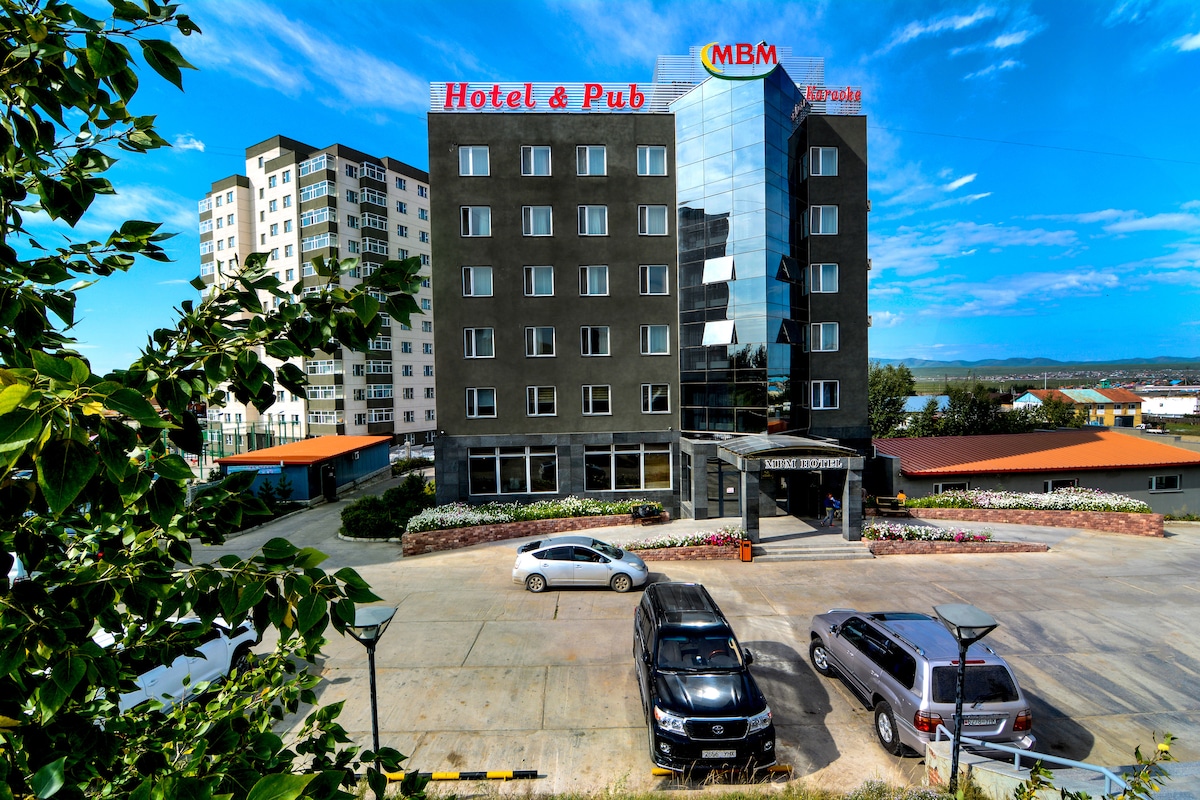 The exterior of the MBM Hotel features a modern five-story building with glass and concrete facades. A vibrant sign displays the hotel's name. A parking area with several vehicles is visible in front, surrounded by landscaped flower beds and greenery under a bright blue sky.