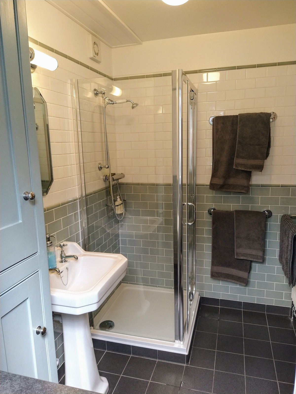 A modern shower room is displayed, featuring a glass shower enclosure and a washbasin with a mirror above. White tiles line the walls, complemented by grey accent tiles. Two grey towels are neatly hung on towel bars, and a dark floor adds contrast to the space.