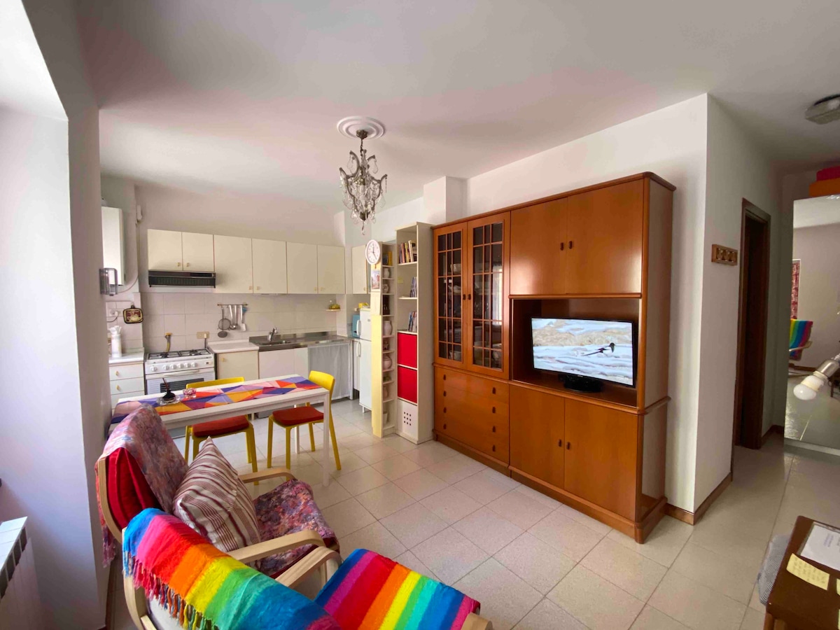 The living area features a combination of colorful furnishings, including a vibrant, striped blanket draped over an armchair. A wooden cabinet houses a flat-screen TV, while a dining table with colorful chairs is positioned nearby. The open kitchen layout includes modern appliances and white cabinetry.