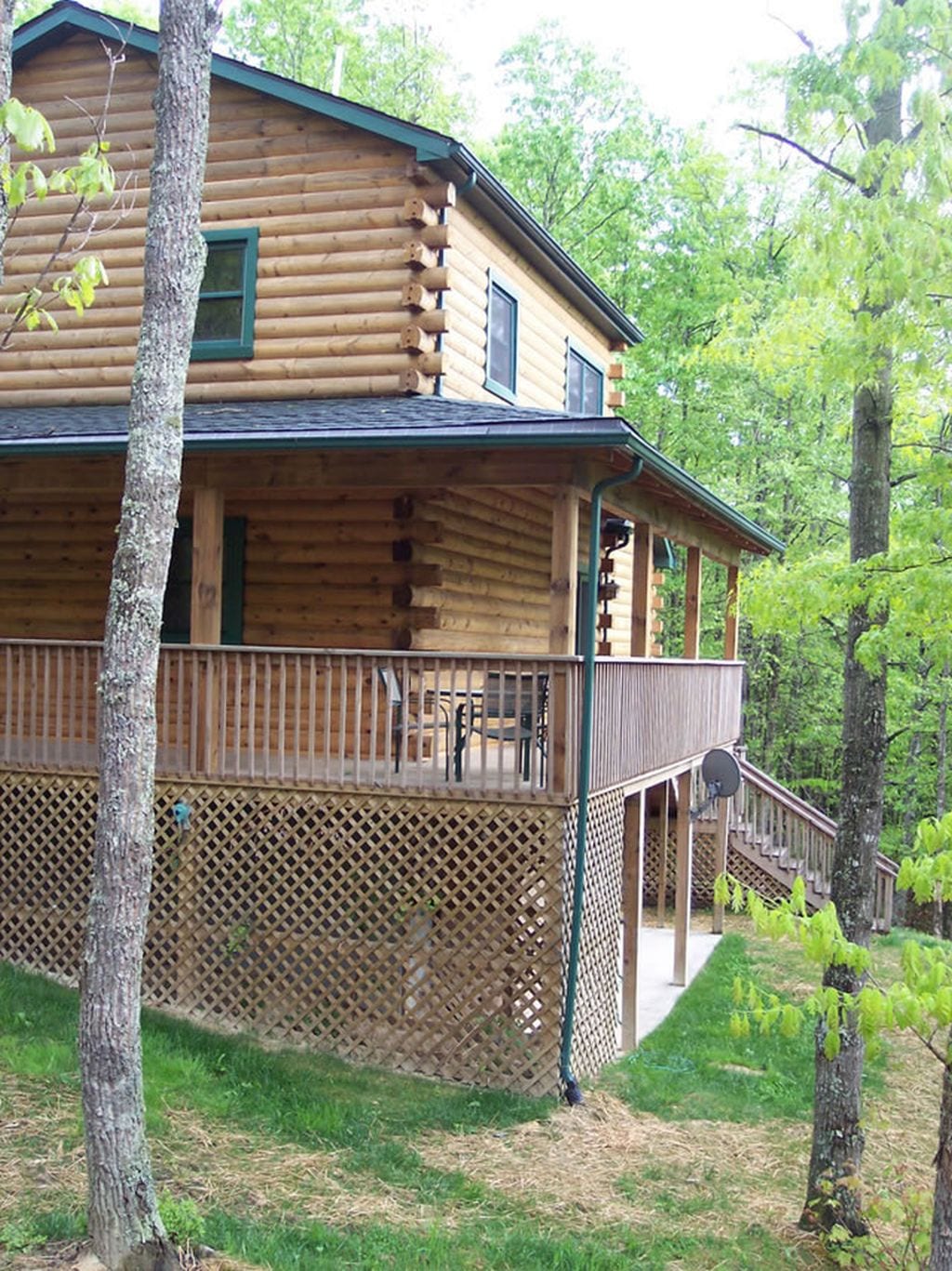 A three-story log cabin is situated within a wooded area, featuring a wrap-around deck with seating. The natural wood structure is complemented by green accents. Surrounding trees provide a serene backdrop, contributing to the secluded ambiance of the property.