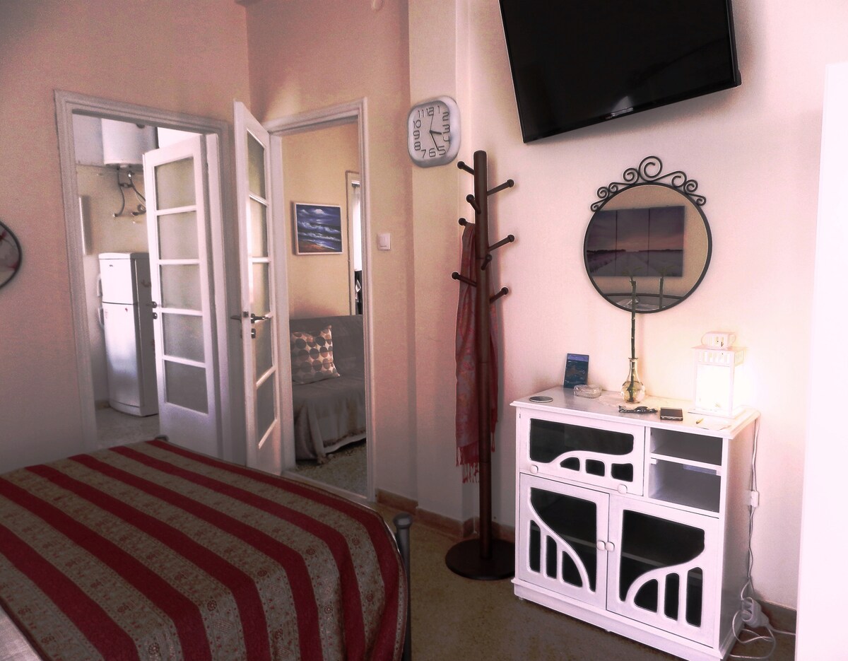 A spacious bedroom is presented, featuring a striped bedspread on a large bed. A wall-mounted TV is visible, along with a mirror and a decorative cabinet. A coat rack stands nearby, and a door leads to a well-lit kitchenette area, enhancing the room's inviting layout.