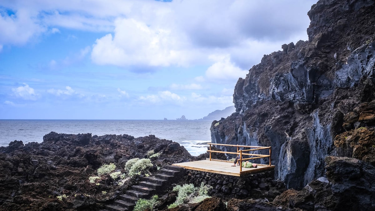 A rocky coastline features a wooden viewing platform overlooking the ocean. Surrounding volcanic rock formations and lush green vegetation frame the scene. The sky is partly cloudy, allowing natural light to illuminate the landscape.