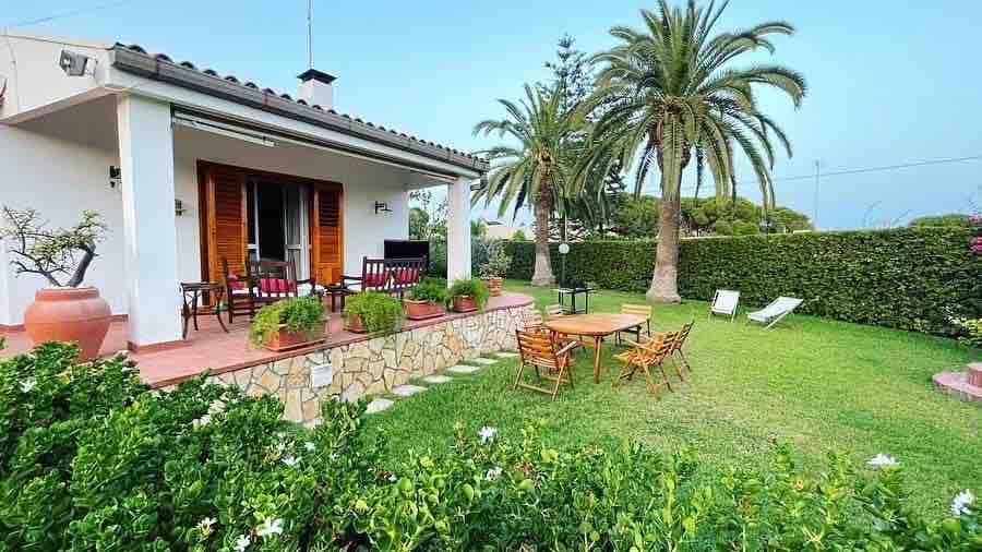 A spacious outdoor area is presented, featuring a well-manicured lawn surrounded by vibrant greenery. Comfortable seating arrangements are visible on the patio, along with a wooden dining table. Nearby, palm trees add to the serene ambiance of this inviting space.