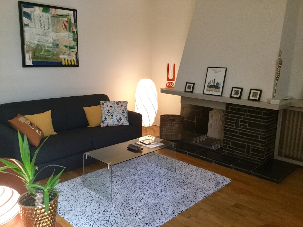A welcoming living area features a dark sofa adorned with decorative cushions, facing a sleek glass coffee table. A soft area rug covers the wooden floor, while art decorates the walls, contributing to an inviting and cozy ambiance.