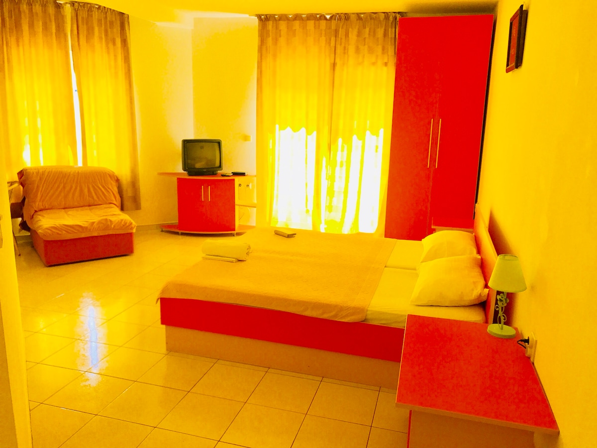 A bright and spacious room features a large bed with light bedding, accented by a small bedside lamp. A sofa bed and a television are also present. Sunlight filters through sheer yellow curtains, illuminating the room's simple design and warm color scheme.
