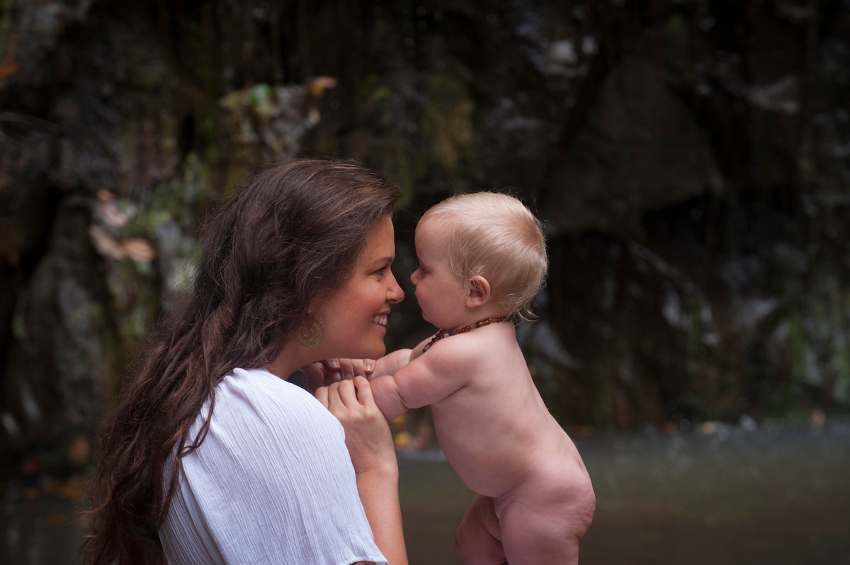 A parent and an infant are engaged near a tranquil water feature. The parent is holding the infant upright, with a rich, natural backdrop of rocks and foliage visible, suggesting a connection to nature.