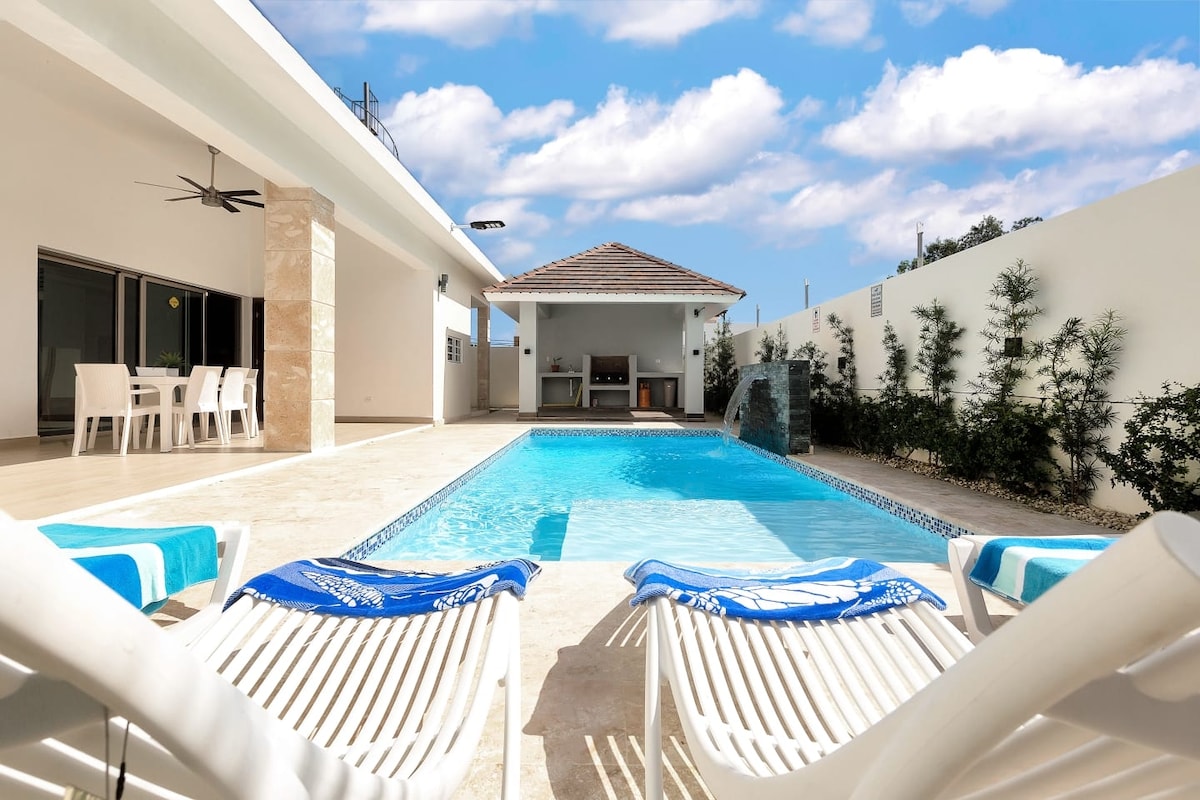 A serene outdoor area features a turquoise pool bordered by light-colored tiles. Two lounge chairs with blue towels are positioned by the pool, and a shaded seating area with a grill is visible in the background. Lush greenery lines the perimeter, creating a tranquil setting.