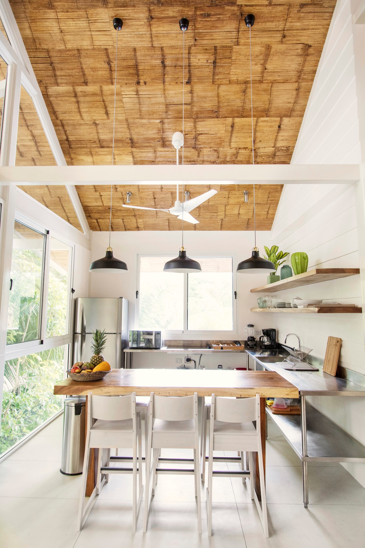 A modern kitchen is displayed with a high thatched roof and large windows allowing natural light. It features sleek stainless-steel appliances, a wooden dining table with white chairs, overhead pendant lighting, and open shelving showcasing cooking essentials and fresh fruits.