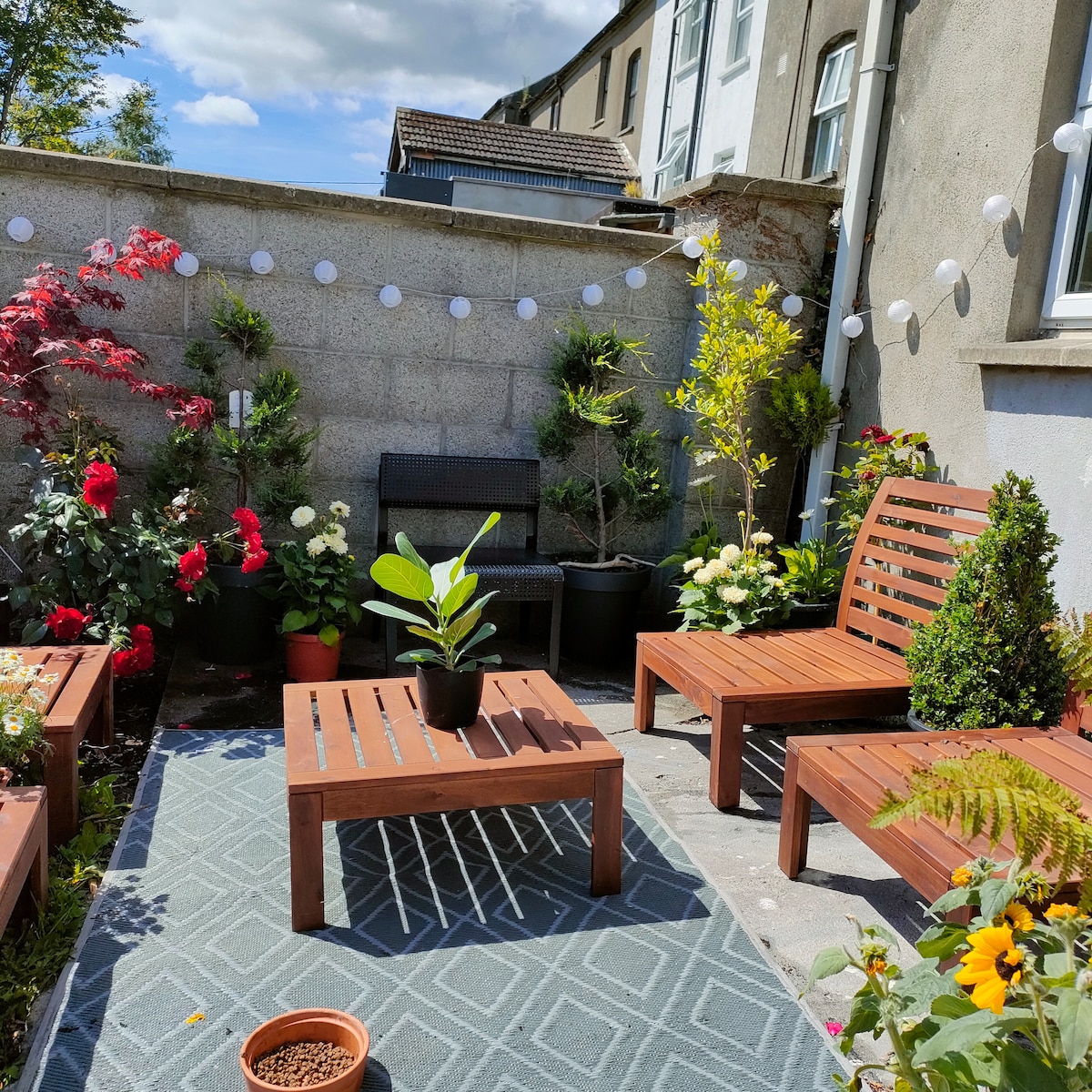 A tranquil outdoor space is presented, featuring wooden seating arranged around a low table. Potted plants and flowers surround the area, adding greenery and color. Decorative string lights are visible along the wall, and a patterned rug is placed on the ground.