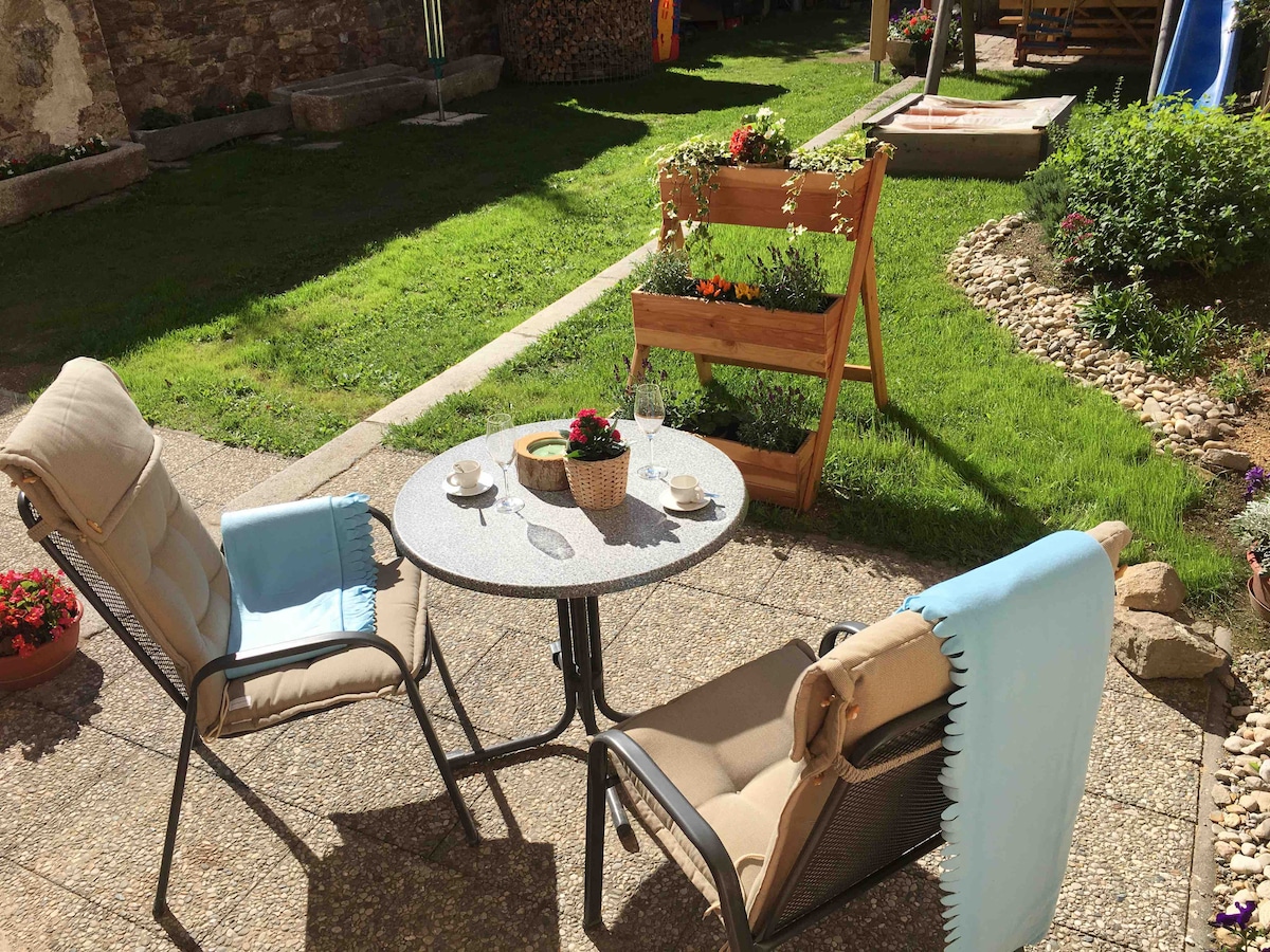 A peaceful outdoor seating area is presented, featuring a small table accompanied by two chairs. Potted flowers add color to the surroundings, while a decorative wooden shelf displays plants. Sunlight illuminates the grassy area, creating a serene atmosphere.