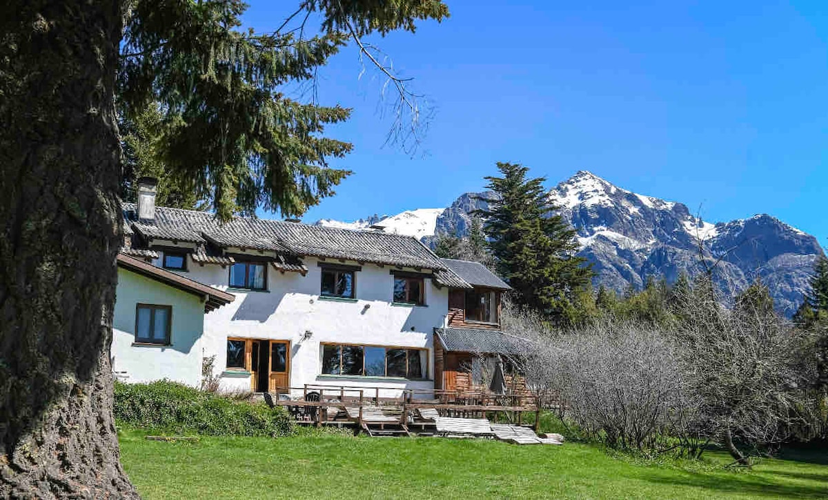 The two-story house features a white exterior with a rustic wooden roof. Large windows allow natural light to fill the interior, while a spacious wooden deck extends towards a lush green lawn. Snow-capped mountains rise in the background under a clear blue sky.