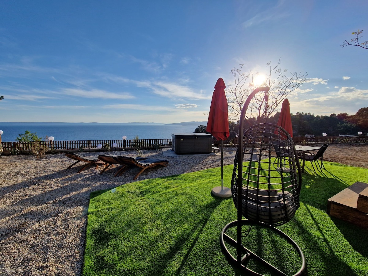 An outdoor space offers a well-maintained lawn area with a hanging chair and sun loungers. Two parasols provide shade against the sun, while the sparkling sea is visible in the background, creating a serene atmosphere.