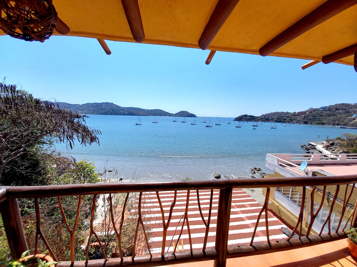 A scenic view is offered from a balcony, revealing tranquil waters and a collection of sailboats anchored in the bay. The shoreline features gentle waves and a landscape of distant hills under a clear blue sky.