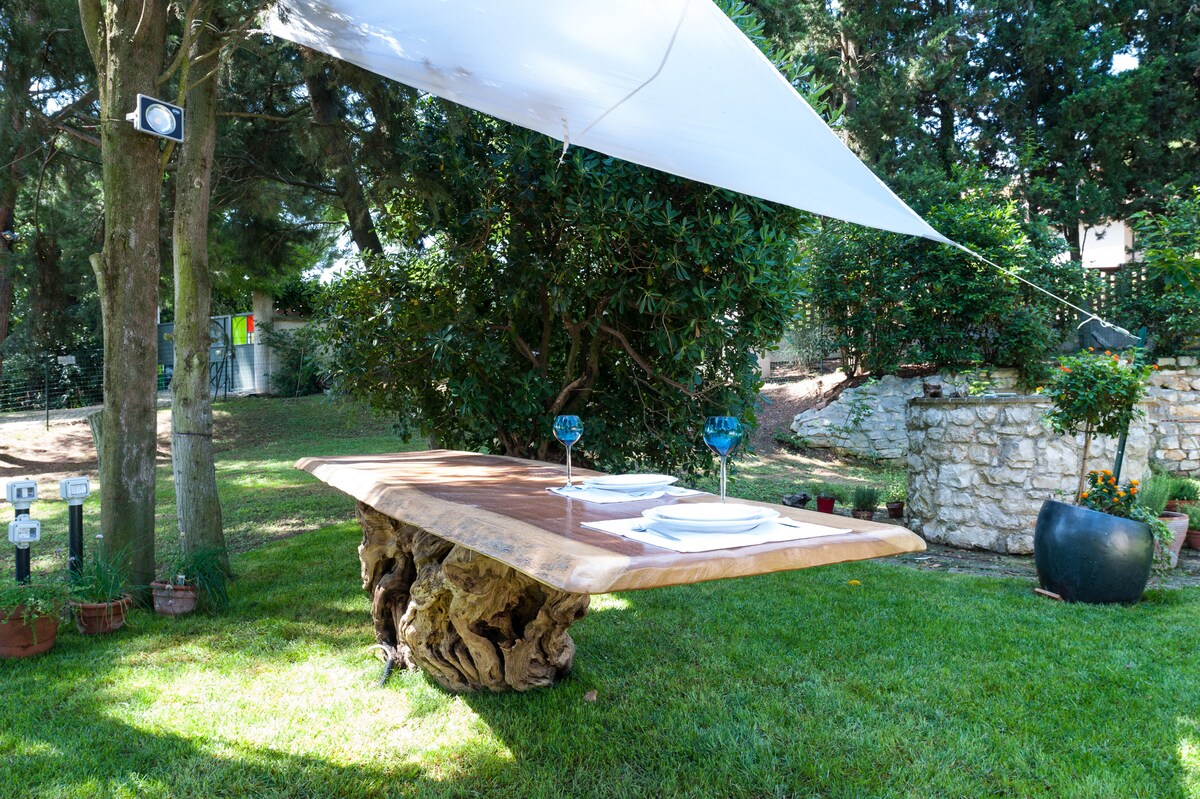 An outdoor dining area features a unique wooden table supported by a sculptural base. Two plates and blue glasses are set on the table, shaded by a large sail. Lush greenery surrounds the space, enhancing the natural ambiance of the garden.