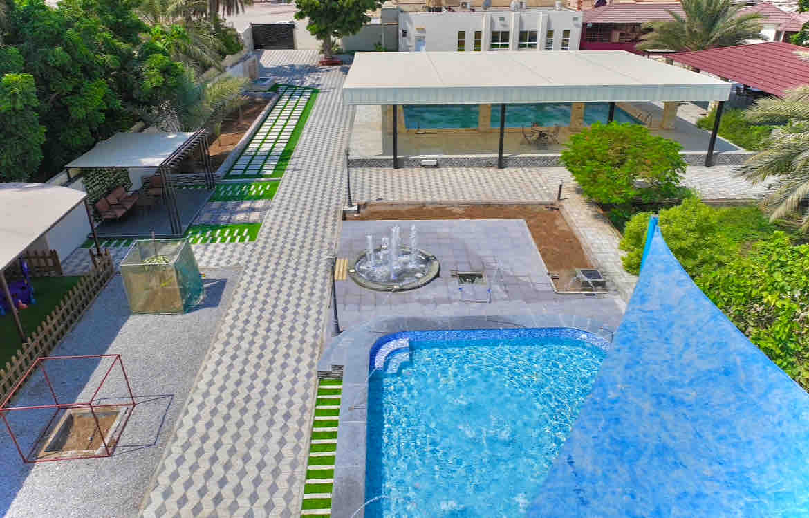 An aerial view reveals a spacious outdoor area featuring a shimmering pool, surrounded by patterned stone paving and green grass. Various seating areas, shaded structures, and a fountain are visible, blending harmoniously into the inviting landscape.