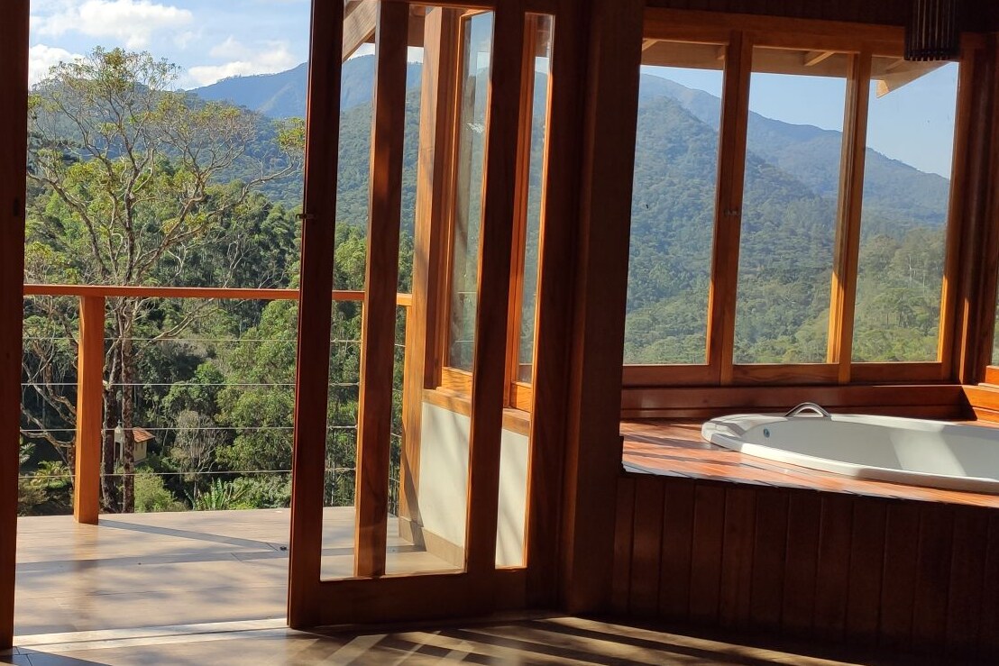 A spacious room is highlighted by large glass doors that open to a wooden deck and scenic mountain views. Natural light fills the space, while a hot tub is positioned nearby, inviting relaxation amid the serene landscape. The wooden finishes add warmth to the interior.