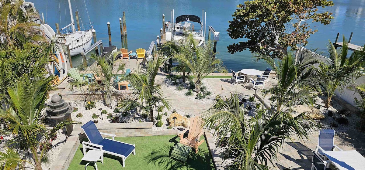 A waterfront backyard is featured, highlighting a spacious area with tropical landscaping. Multiple seating options include lounge chairs and tables. A dock for boating and fishing is visible, along with boats moored nearby, creating a serene atmosphere by the water.