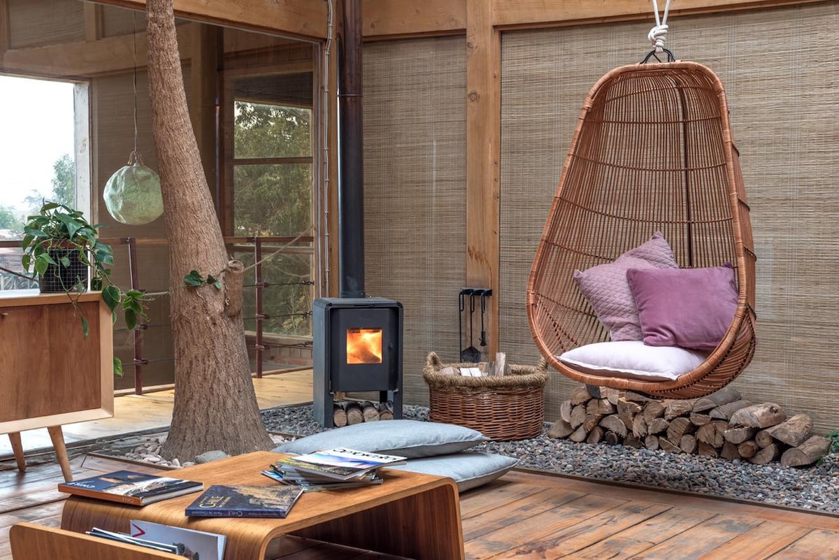A cozy seating area features a suspended wicker chair adorned with soft cushions. A small wood-burning stove provides warmth, positioned near a stack of firewood. The space has natural finishes, including wooden beams and a tree, enhancing the overall charm.