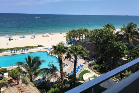 A view from a balcony showcases a sandy beach and ocean waves under a clear sky. A swimming pool is visible in the foreground, surrounded by lounge chairs and palm trees. Beachgoers can be seen enjoying the sunny day on the shore.