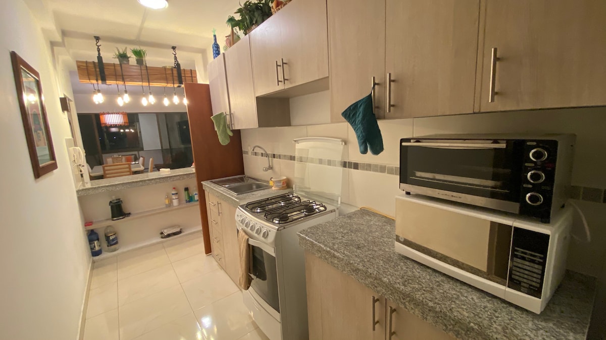 A modern kitchen is showcased with light wood cabinetry and marble countertops. The kitchen is equipped with a gas stove, oven, microwave, and a silver refrigerator. Colorful oven mitts hang from the cabinetry while stylish pendant lights illuminate the space.