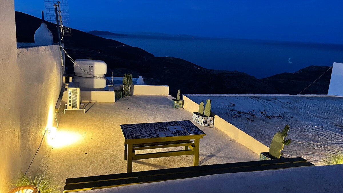 A serene terrace is illuminated by warm lighting, showcasing a low table surrounded by decorative planters. The expansive space offers views of the ocean, with distant hills visible under the evening sky. Potted plants are placed strategically along the edge.